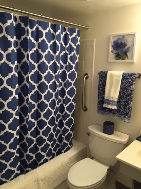 A bathroom with a white toilet, a blue and white patterned shower curtain, a white towel hanging on a towel rack with a blue patterned towel underneath, a small blue container on the toilet tank, a grab bar on the wall next to the toilet, and a framed floral artwork above the towel rack.