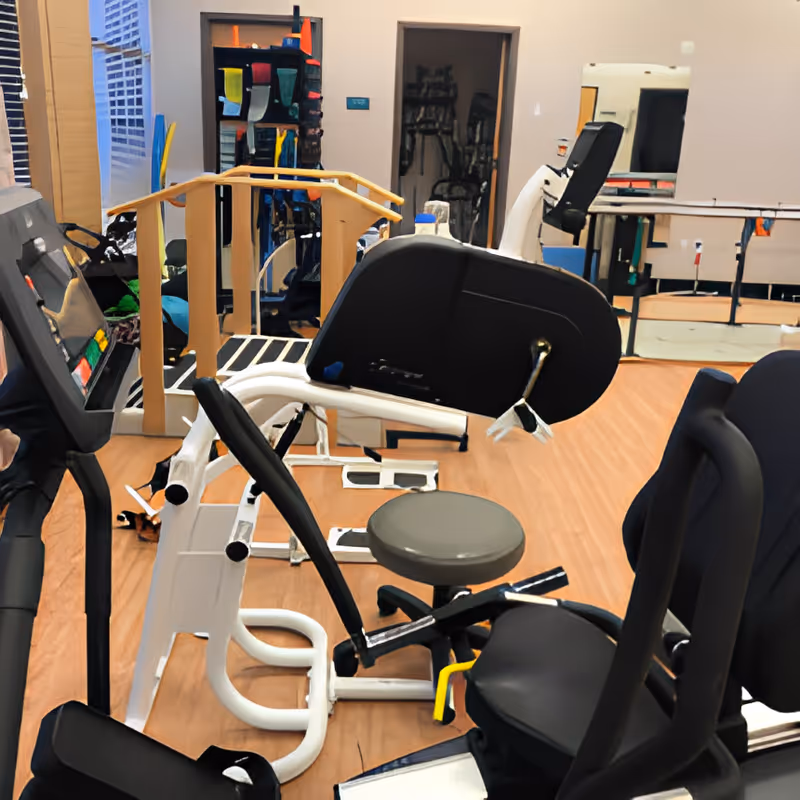 A physical therapy/exercise room with various gym machines, a treadmill, parallel bars and seating on wood flooring.