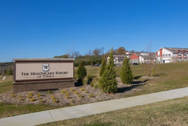 Outdoor view of The Healthcare Resort of Topeka sign with landscaped bushes and grass in front, and the facility building visible in the background under a clear blue sky.