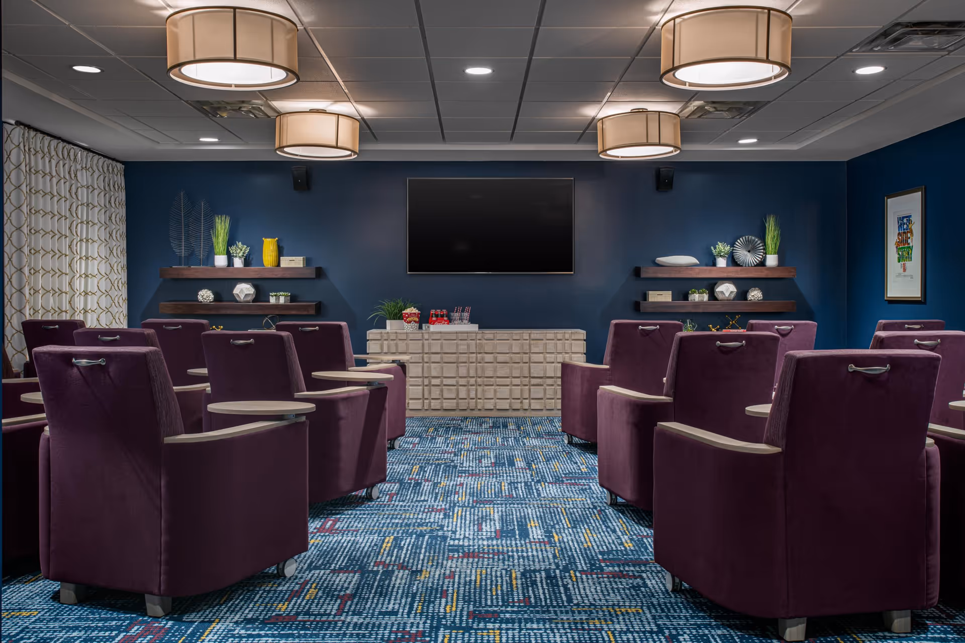 A cozy media room with rows of purple armchairs equipped with small attached tables, facing a large flat-screen TV mounted on a dark blue wall. The room features patterned blue carpet, modern ceiling lights, decorative shelves with plants and ornaments, and a sideboard beneath the TV.