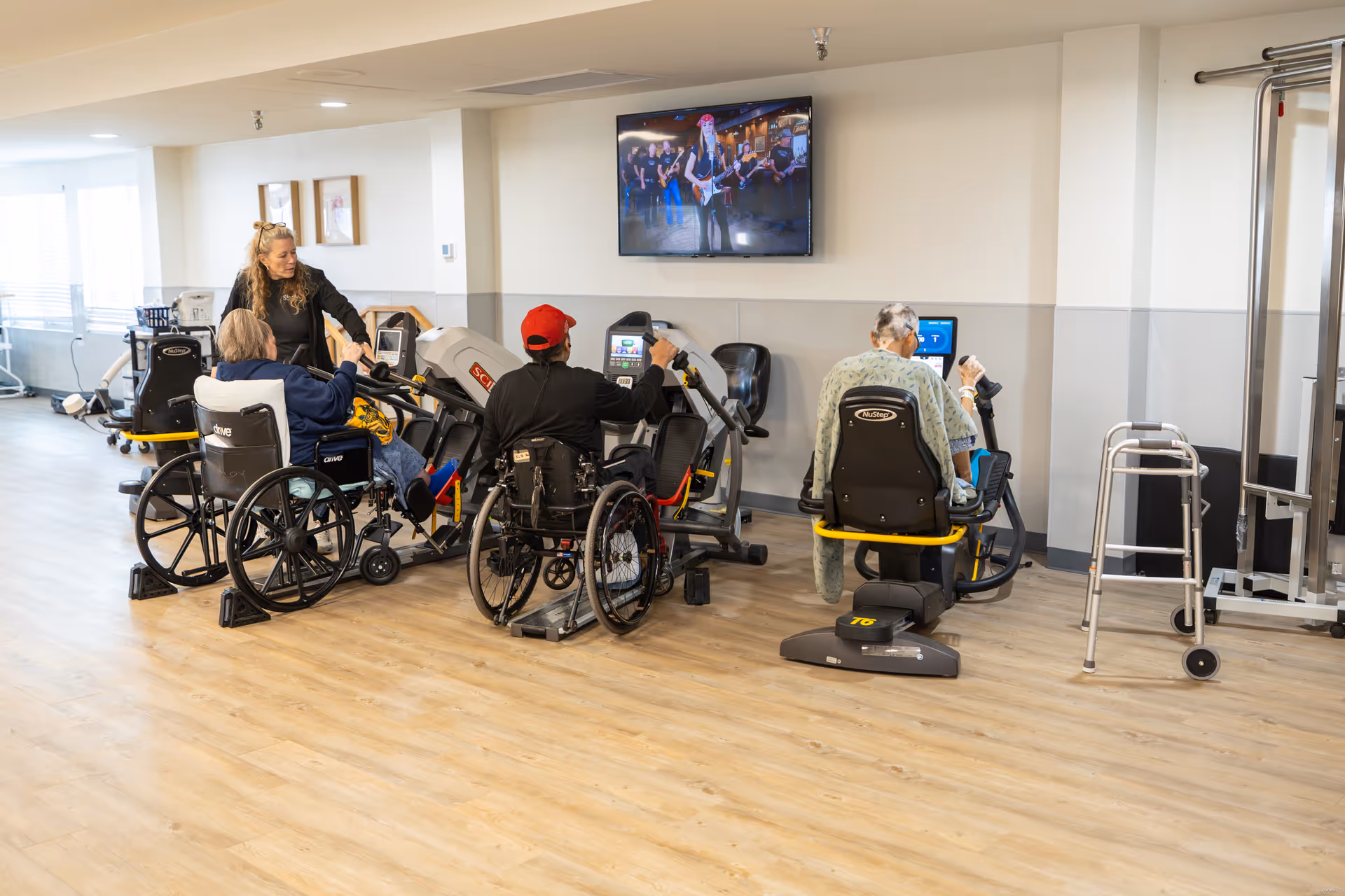 A rehabilitation room with three elderly individuals using exercise equipment, two in wheelchairs and one seated on a recumbent exercise bike. A caregiver is assisting one of the individuals. A television mounted on the wall shows a music performance. The room has light-colored walls and wood flooring, with additional exercise equipment and a walker visible.