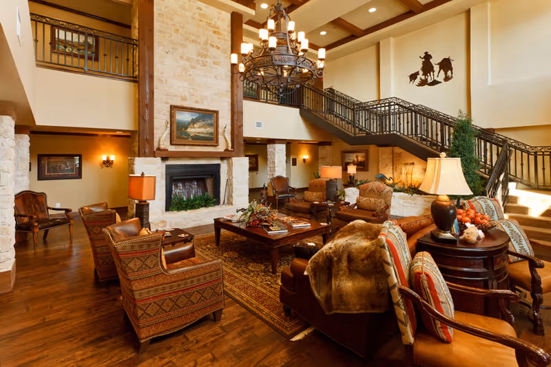 A spacious and warmly decorated living room area with a large stone fireplace, wooden beams, and a chandelier hanging from the ceiling. The room features comfortable upholstered chairs and sofas arranged around a wooden coffee table on a patterned rug. There is a staircase with wrought iron railings leading to an upper level, and various lamps and framed artwork adorn the walls.