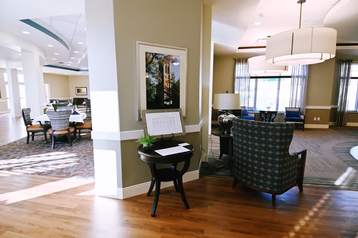 A bright, spacious interior area of a senior living facility featuring a seating area with patterned armchairs, a side table with a lamp, and large windows with sheer curtains. Adjacent to this is a dining area with round tables and chairs, carpeted flooring, and decorative ceiling details. A small round table with a plant and framed pictures is positioned against a column in the center.