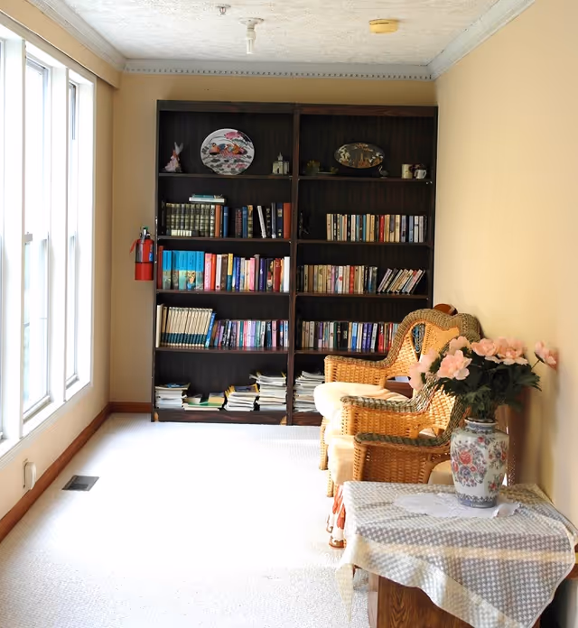 A cozy interior corner with a large dark wooden bookshelf filled with books and decorative plates. Next to the bookshelf are two wicker chairs with cushions. In the foreground, there is a small table covered with a patterned cloth and a vase with pink flowers. Large windows on the left side let in natural light, illuminating the beige walls and light carpeted floor.