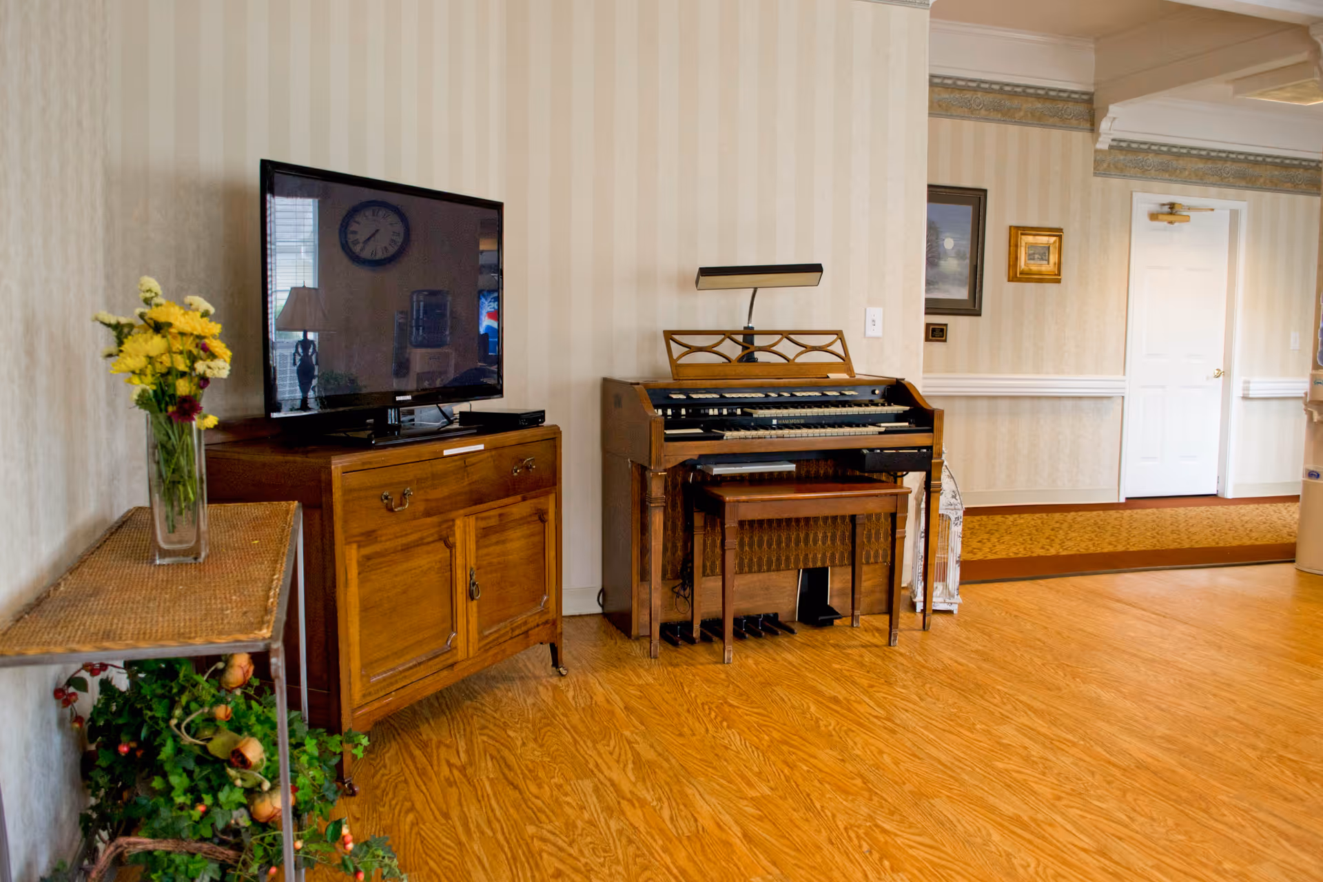 Interior room with wooden flooring featuring a wooden cabinet with a flat-screen TV on top, a small table with a vase of yellow and red flowers, and an electric organ with a bench. The walls have light-colored striped wallpaper, and there are framed pictures and a white door in the background.