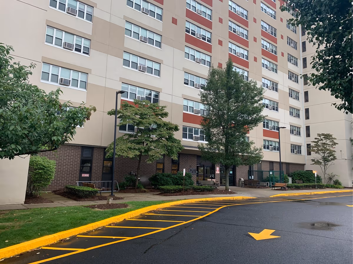 Exterior view of a multi-story residential building with beige and red accents, several windows with air conditioning units, trees and shrubs in front, benches near the entrance, and a wet asphalt driveway with yellow painted lines and an arrow.
