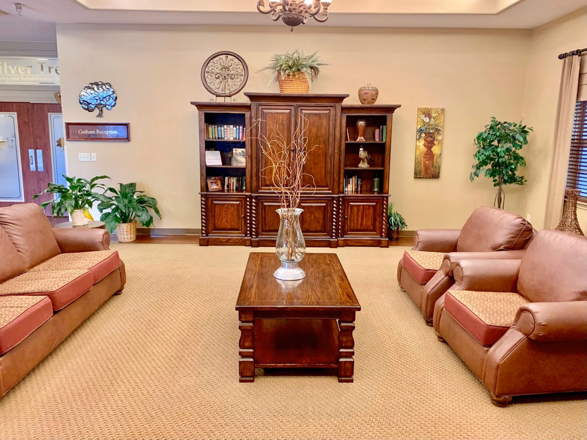 A cozy living room area in a nursing and rehabilitation facility featuring two brown leather sofas and a matching armchair arranged around a wooden coffee table with a glass vase holding decorative branches. Behind the seating area is a large wooden cabinet with shelves holding books and decorative items. The room is decorated with plants, a wall clock, and artwork, with beige walls and carpeted flooring.