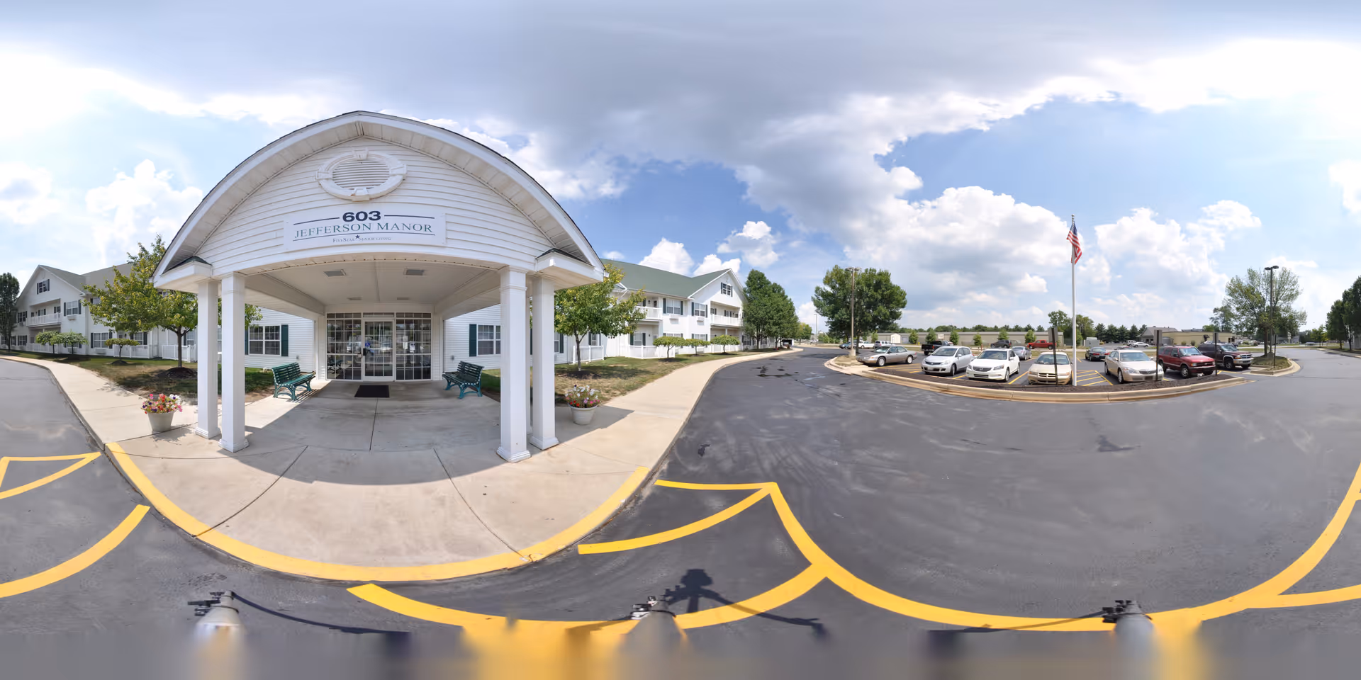 Front entrance of Jefferson Manor with a covered porte-cochere, benches, and an adjacent parking lot under a partly cloudy sky.