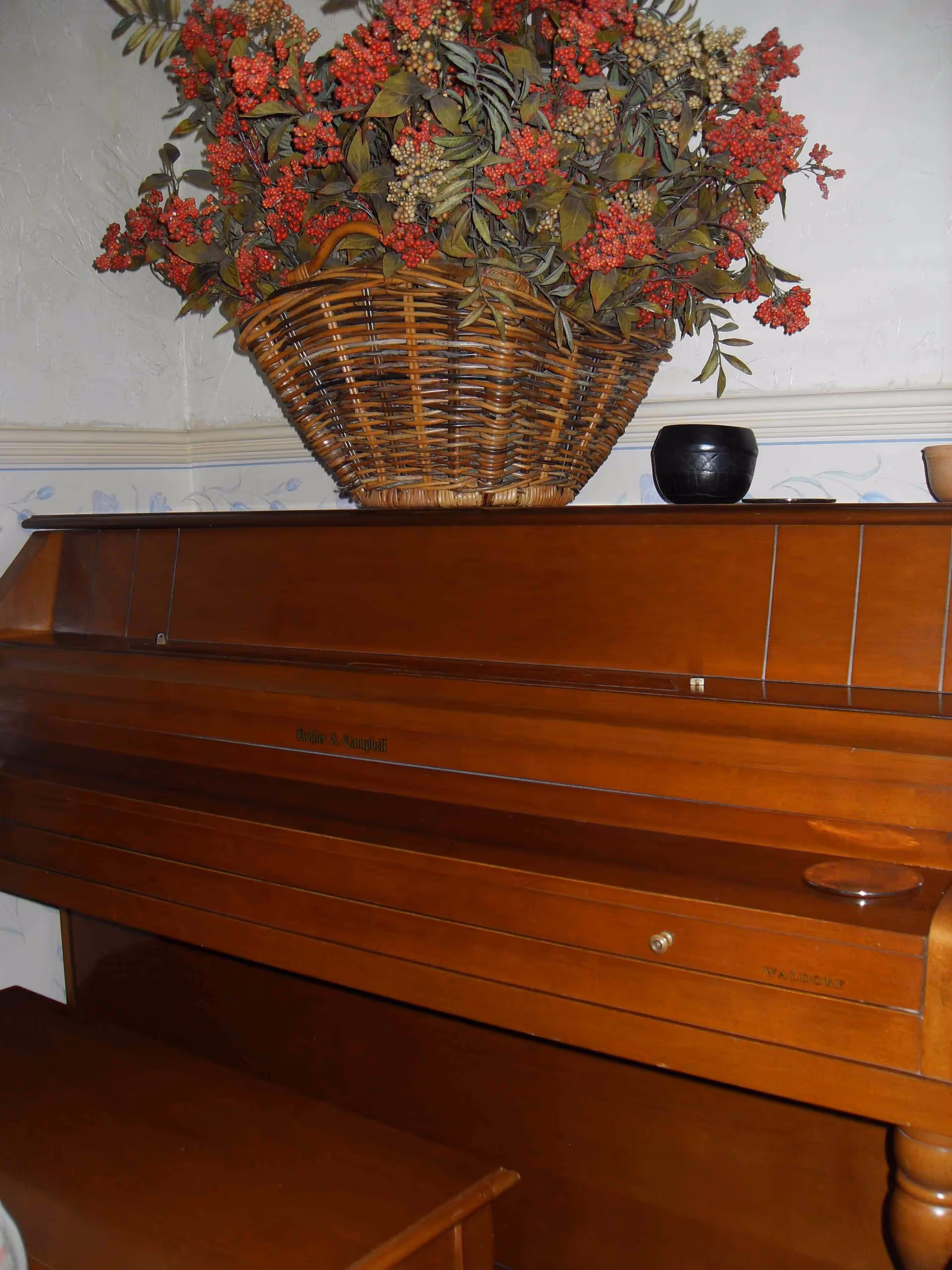 A wooden upright piano with a large wicker basket filled with artificial red and beige berries and green leaves placed on top. There is also a small black pot and a small brown pot on the piano. The background shows a light-colored wall with a floral border wallpaper.