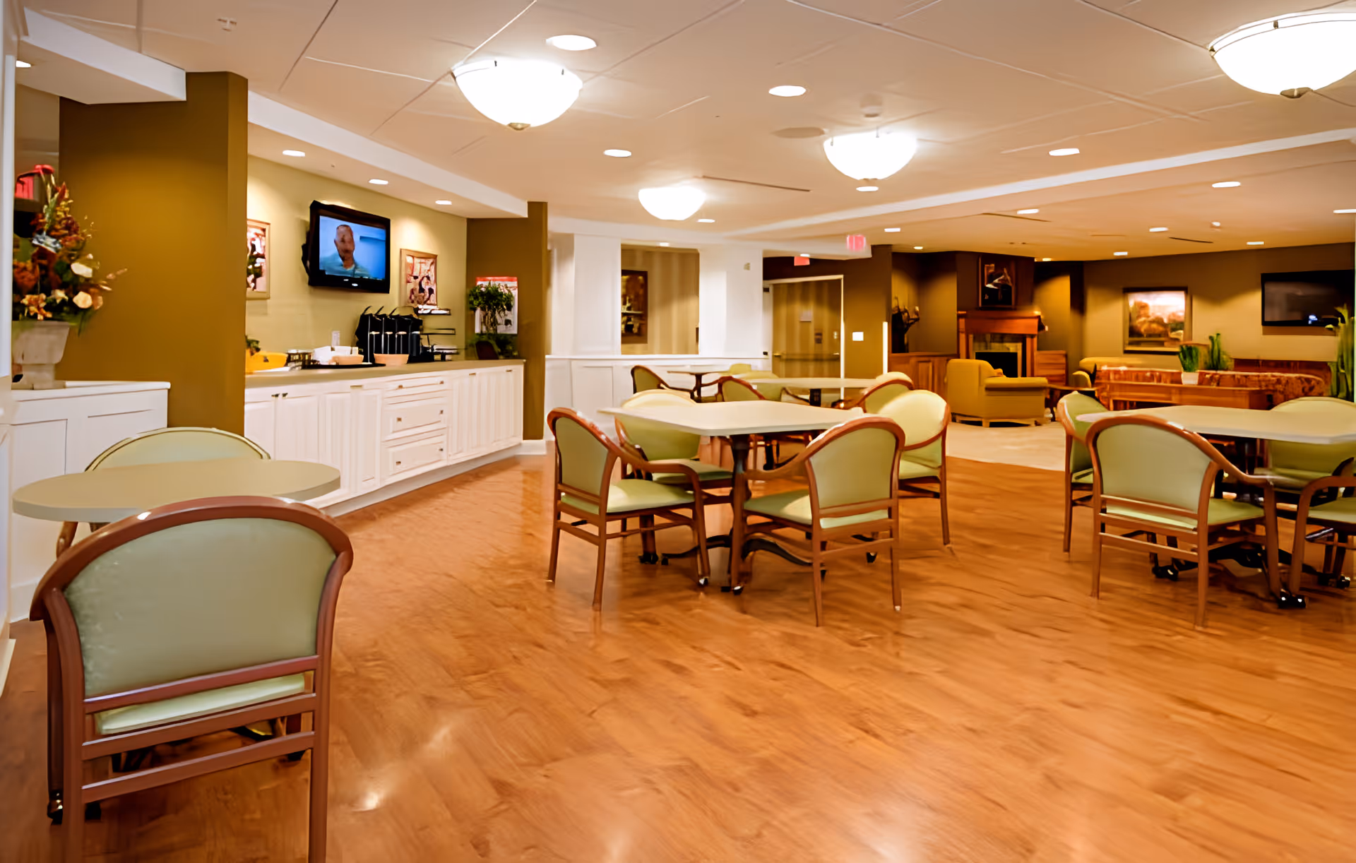 Spacious assisted-living dining and lounge area with round tables and green-cushioned chairs, a serving counter, and wood flooring.