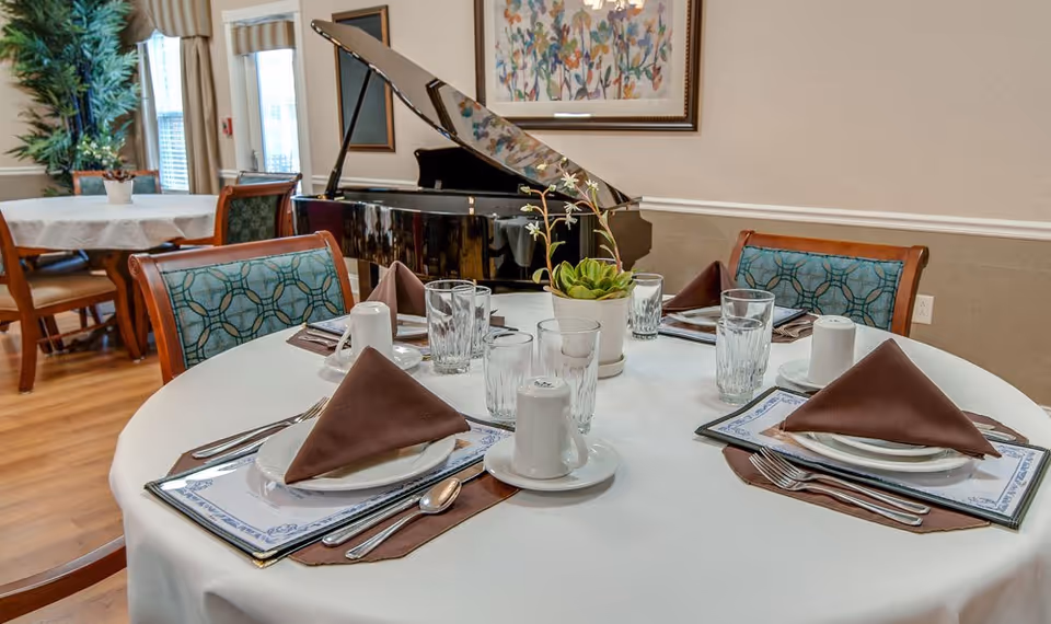 A round dining table set for four with white tablecloth, brown folded napkins, plates, glasses, and silverware. In the background, there is a black grand piano, framed artwork on the wall, and another dining table with chairs near windows with curtains.