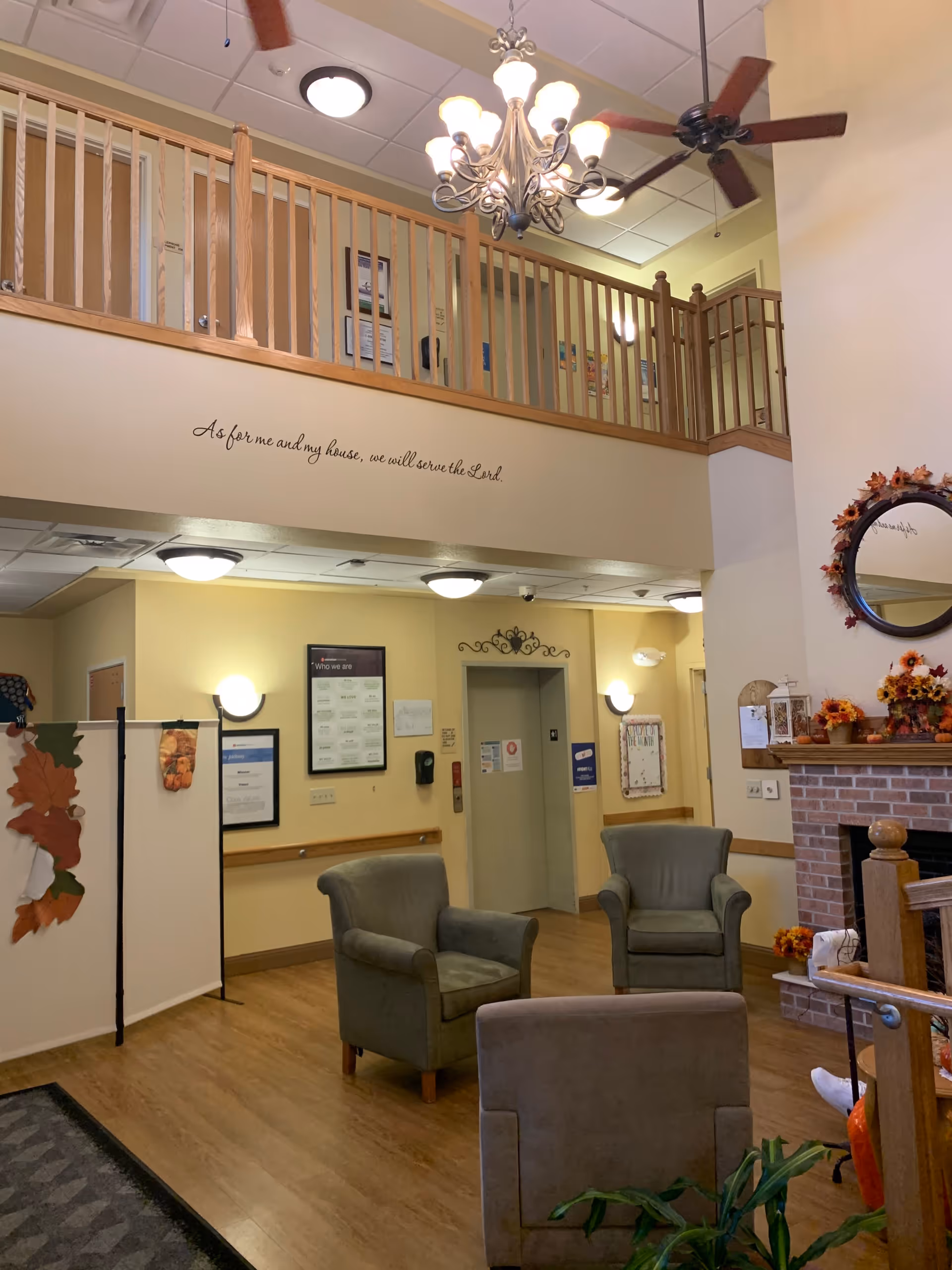 A cozy interior common area with three armchairs arranged on a wooden floor near a brick fireplace decorated with autumn-themed flowers and a round mirror. Above, there is a wooden railing balcony with ceiling fans and a chandelier. The wall features a quote that reads, 'As for me and my house, we will serve the Lord.' There is an elevator and various framed notices on the walls.