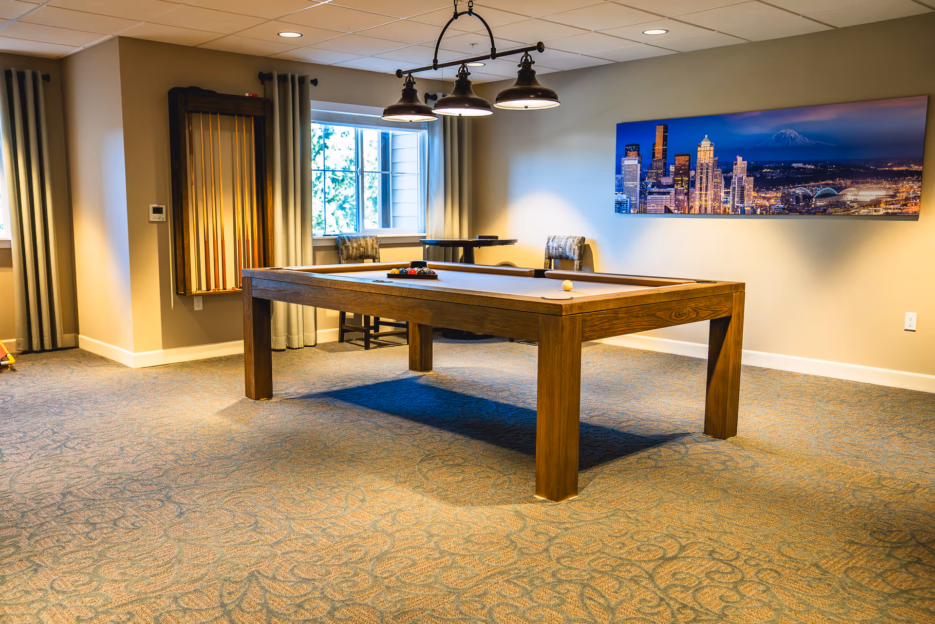A well-lit game room with a wooden pool table in the center, a rack of pool cues mounted on the wall, two chairs near a window with curtains, and a large panoramic cityscape photograph hanging on the wall.