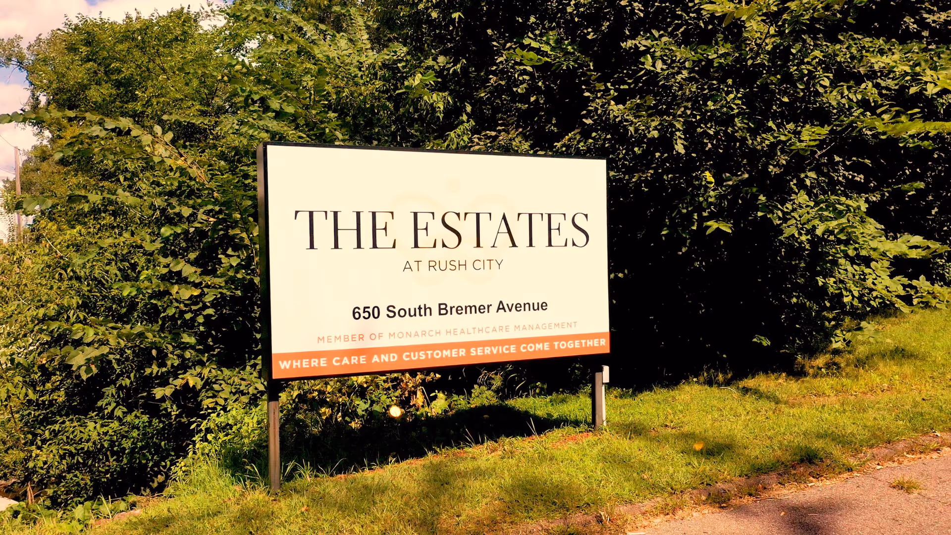 A large outdoor sign for The Estates at Rush City located on 650 South Bremer Avenue, surrounded by green bushes and grass under a partly cloudy sky.