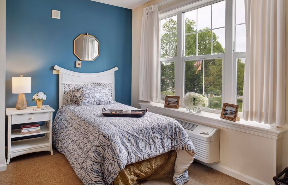 A cozy bedroom with a single bed covered in a patterned blue and white comforter. There is a white wicker headboard and a matching nightstand with a lamp and a small flower pot. A small octagonal mirror hangs on the blue accent wall above the bed. Large windows with white curtains let in natural light and show green trees and an American flag outside. The windowsill holds two framed photos and a vase with white flowers.