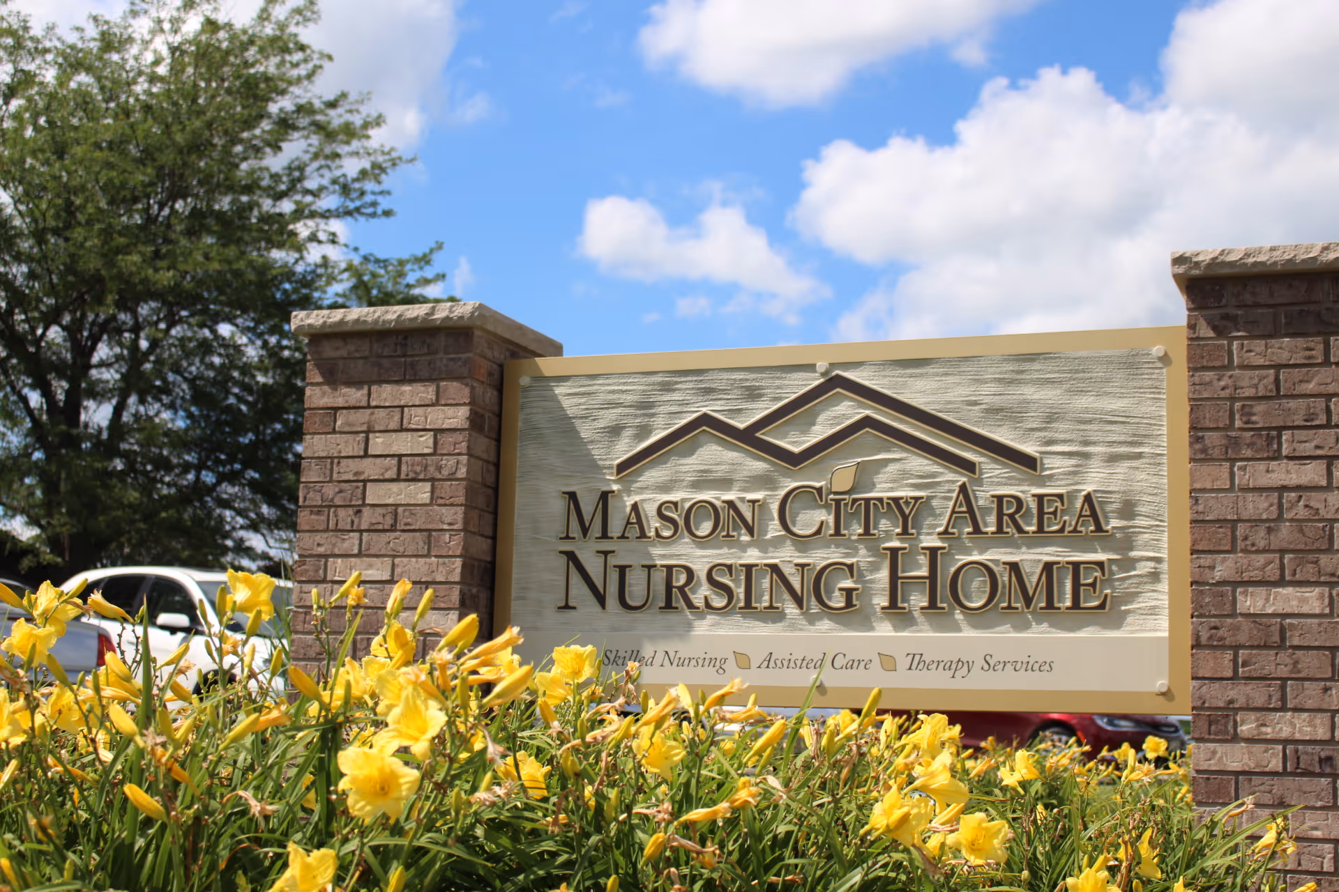 Entrance sign reading 'Mason City Area Nursing Home' with yellow flowers, brick pillars, and a blue sky background.