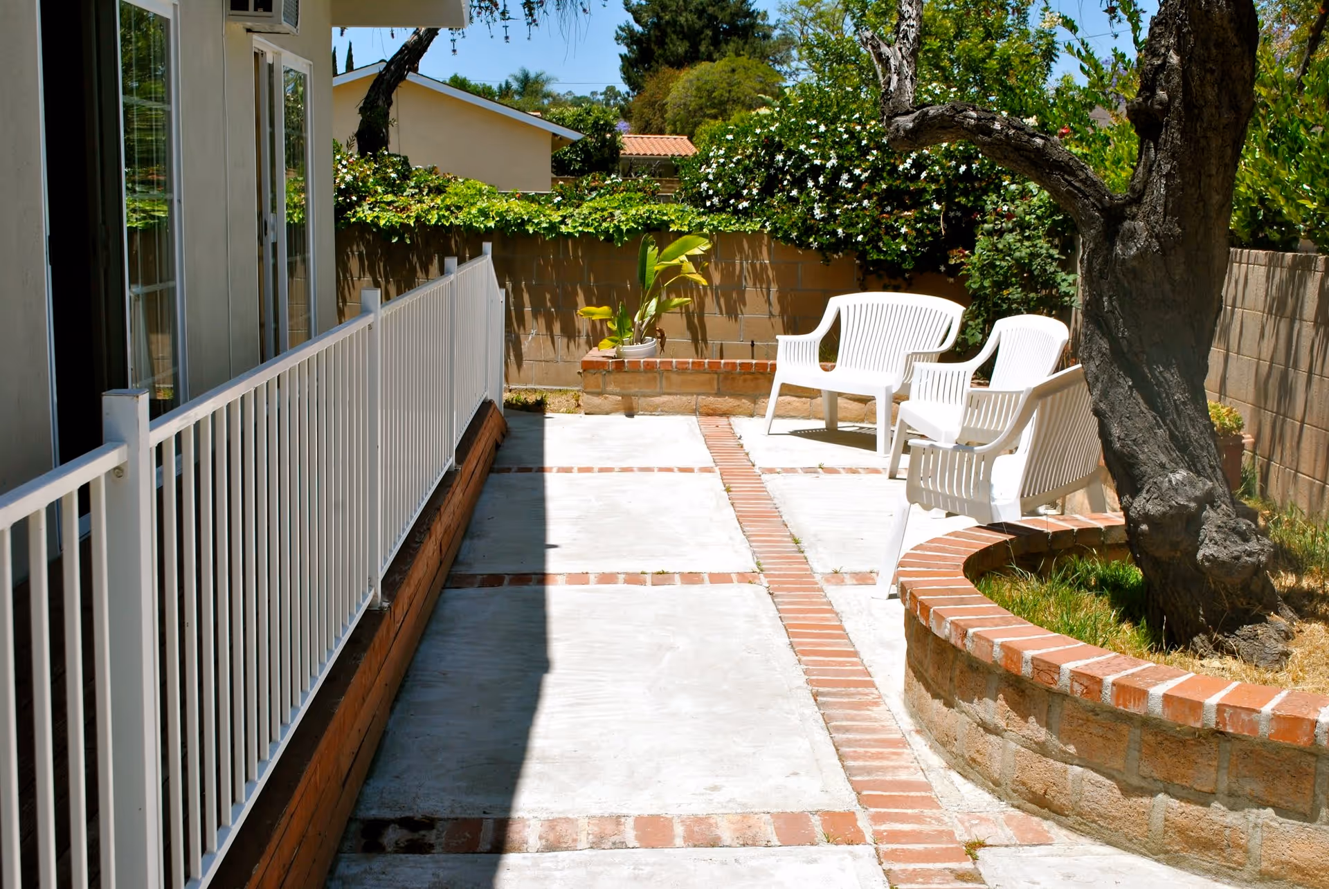 Outdoor patio area with white plastic chairs arranged near a curved brick planter containing a tree. The patio has a concrete floor with brick accents and is enclosed by a low brick wall with greenery and bushes behind it. A white metal railing runs along the left side adjacent to a building with sliding glass doors.