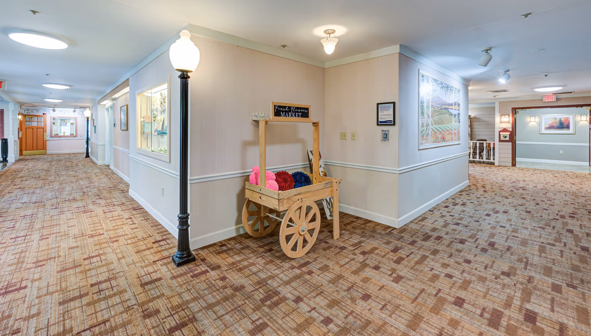 A wide hallway in a senior living facility with patterned carpet and beige walls. There are decorative streetlamp-style lights along the hallway and a wooden cart labeled 'Fresh Flowers Market' holding colorful yarn or fabric. The hallway has framed artwork on the walls and leads to other rooms with visible doorways.