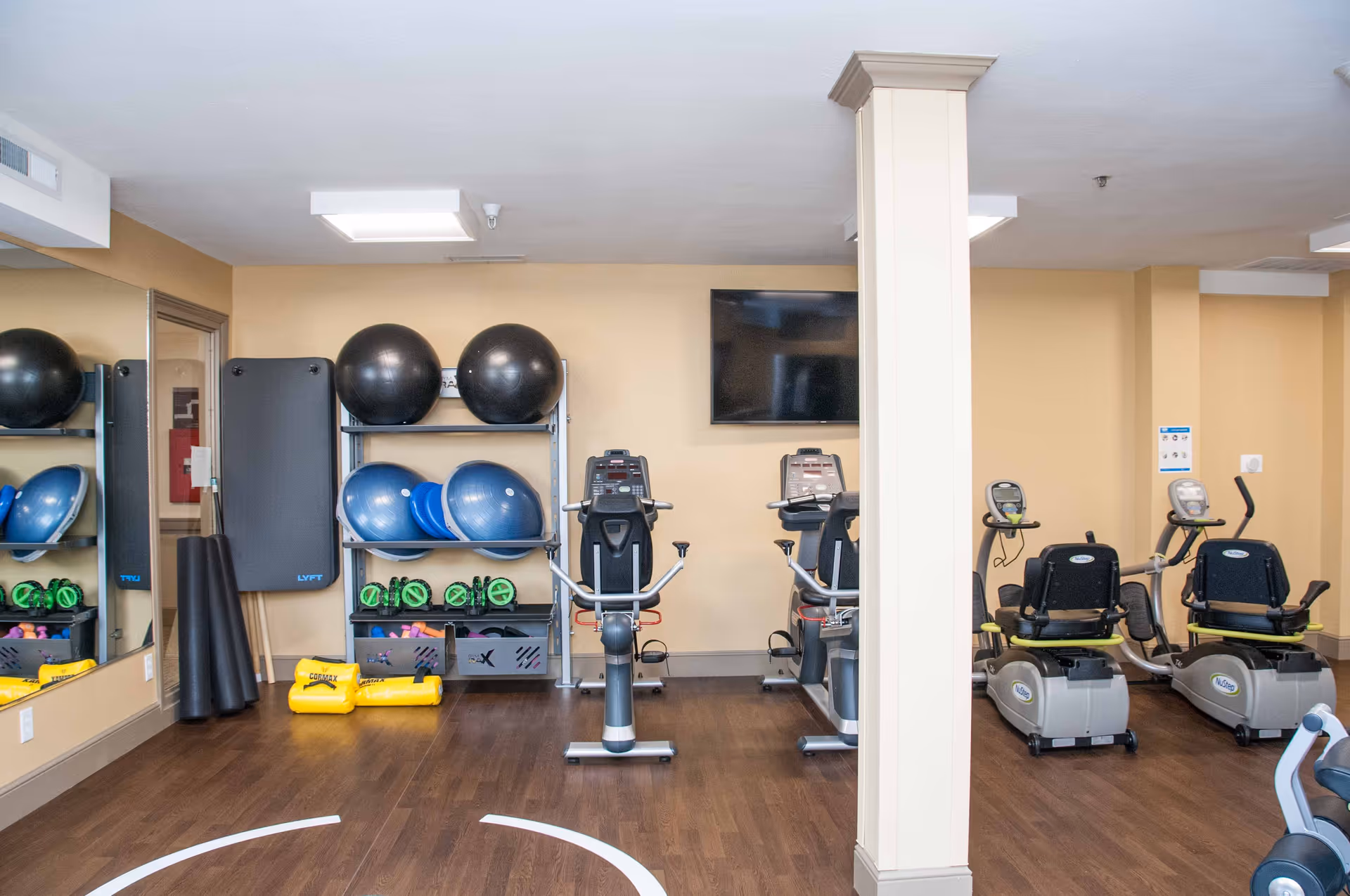 A fitness room with exercise equipment including stationary bikes, recumbent bikes, exercise balls, resistance bands, and yoga mats. There is a large mirror on the left wall and a flat-screen TV mounted on the beige wall in the center. The floor is wooden and the room is well-lit with ceiling lights.