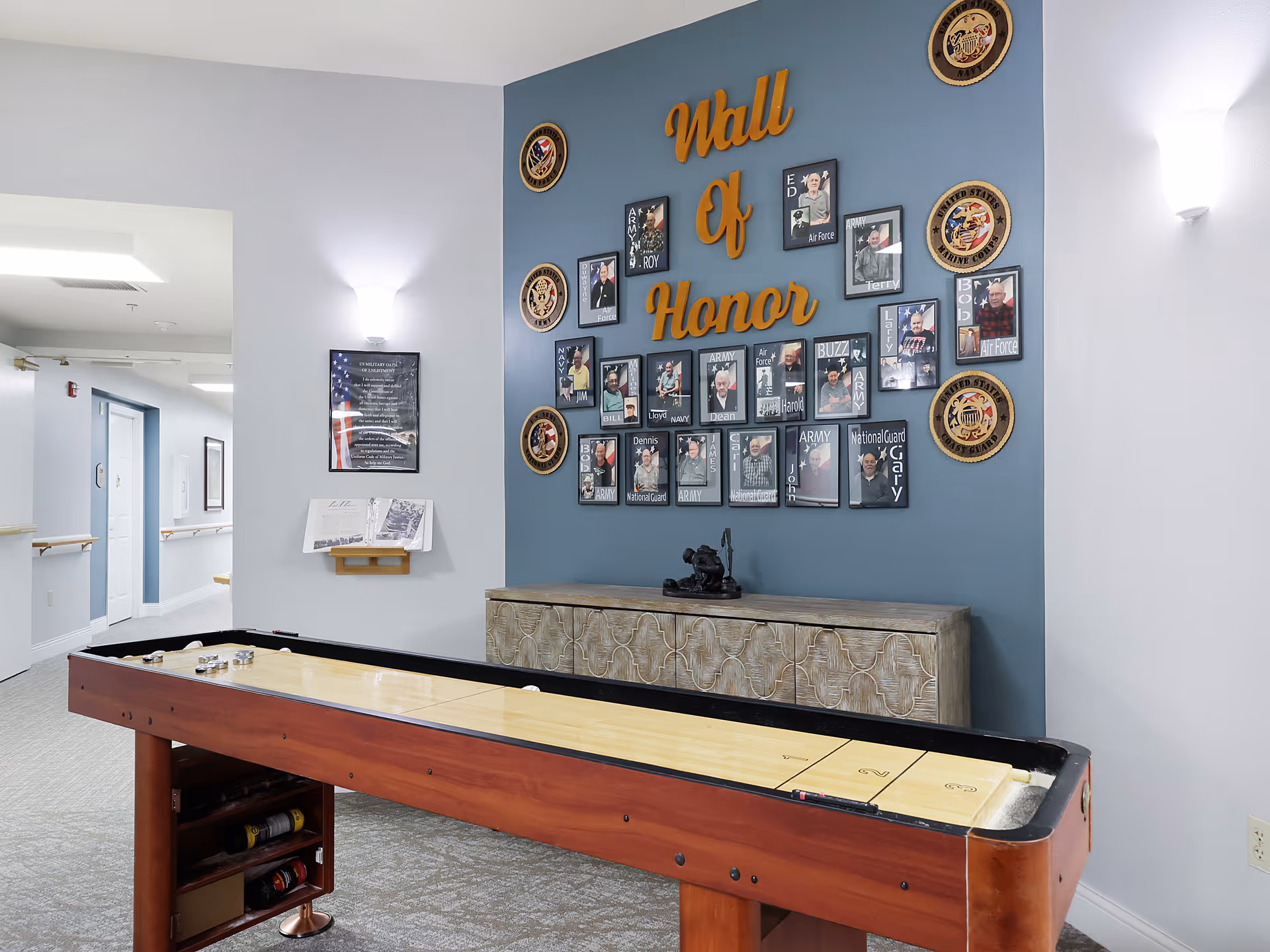 Interior view of a senior living facility hallway featuring a shuffleboard table in the foreground and a 'Wall of Honor' display on a blue accent wall. The display includes framed photos and plaques honoring veterans, with decorative military insignias. The hallway is well-lit with wall sconces and has light-colored walls and carpeted flooring.