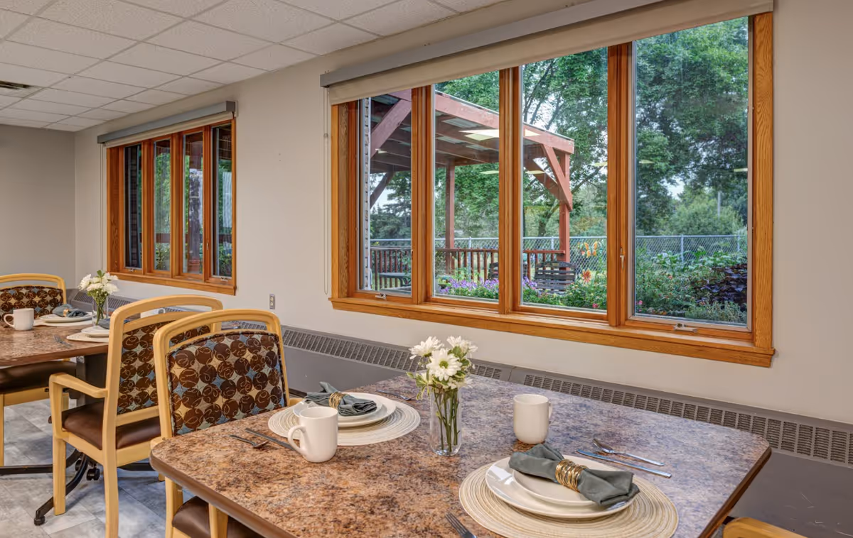 Dining room with set tables and chairs beside large windows overlooking a garden pergola.