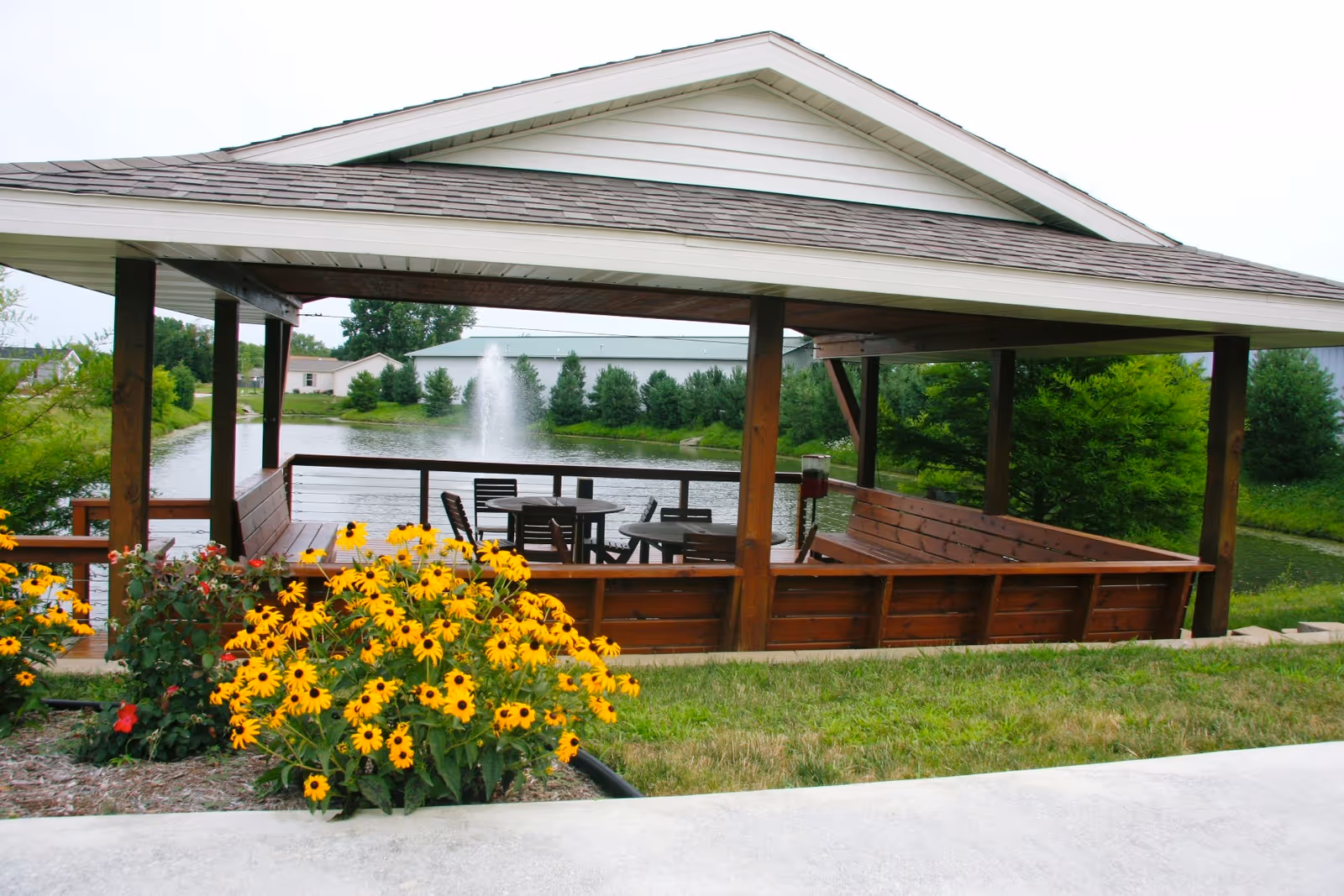 A wooden gazebo with a shingled roof overlooking a pond with a water fountain. Inside the gazebo are round tables and chairs. In the foreground, there are yellow flowers and green grass surrounding the area.