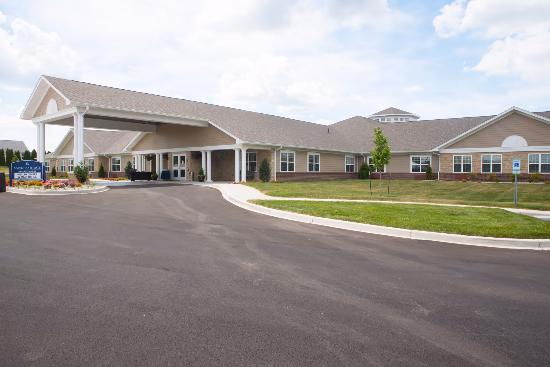 Front entrance and covered drop-off of a single-story senior living campus building with a driveway and landscaped lawn.