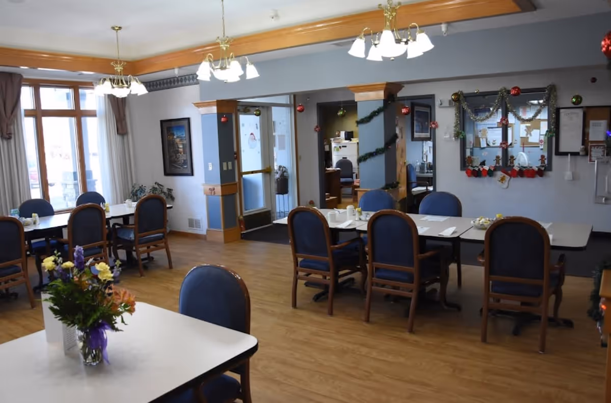A dining room in Cedar Vale Assisted Living with several tables and blue cushioned chairs. The room has wooden flooring, large windows with curtains, and ceiling lights. There are holiday decorations such as garlands and ornaments hanging on the walls and mirrors. A vase with flowers is placed on one of the tables.