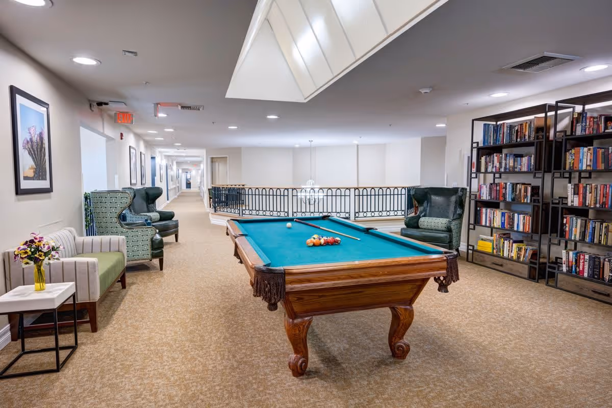 A spacious indoor common area featuring a pool table with balls and cues arranged on it. Surrounding the pool table are comfortable armchairs and a small sofa with a side table holding a vase of flowers. On the right side, there are tall bookshelves filled with books. The area is well-lit with ceiling lights and has a beige carpeted floor. A hallway extends in the background with framed pictures on the walls.