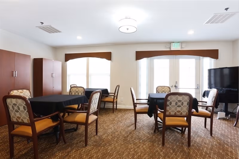 A bright communal dining room with several tables covered in black tablecloths and upholstered chairs, a TV in the corner, and double doors with windows.