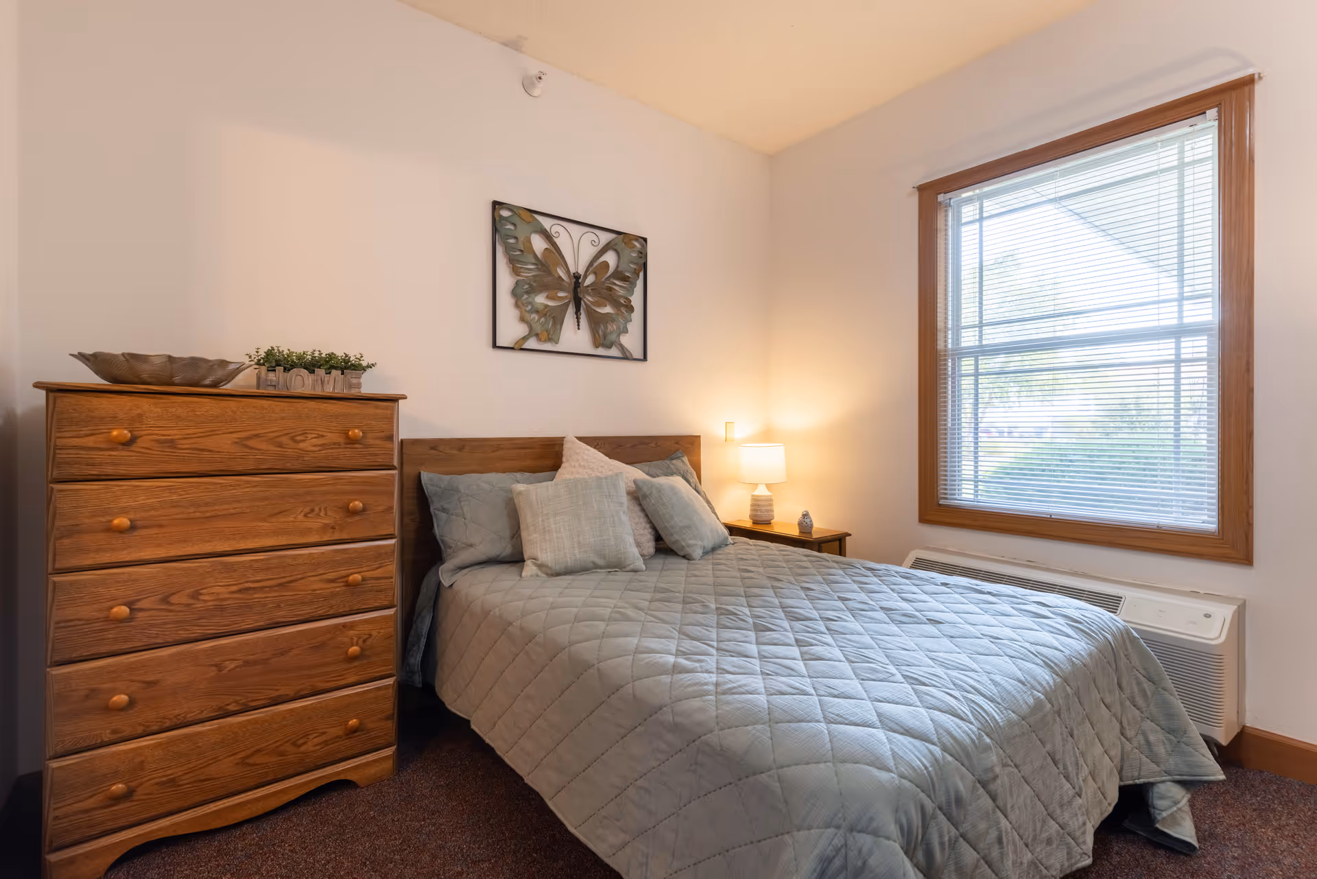 A cozy furnished bedroom with a made bed, wooden dresser, nightstand with lamp, window with blinds, and butterfly wall art.