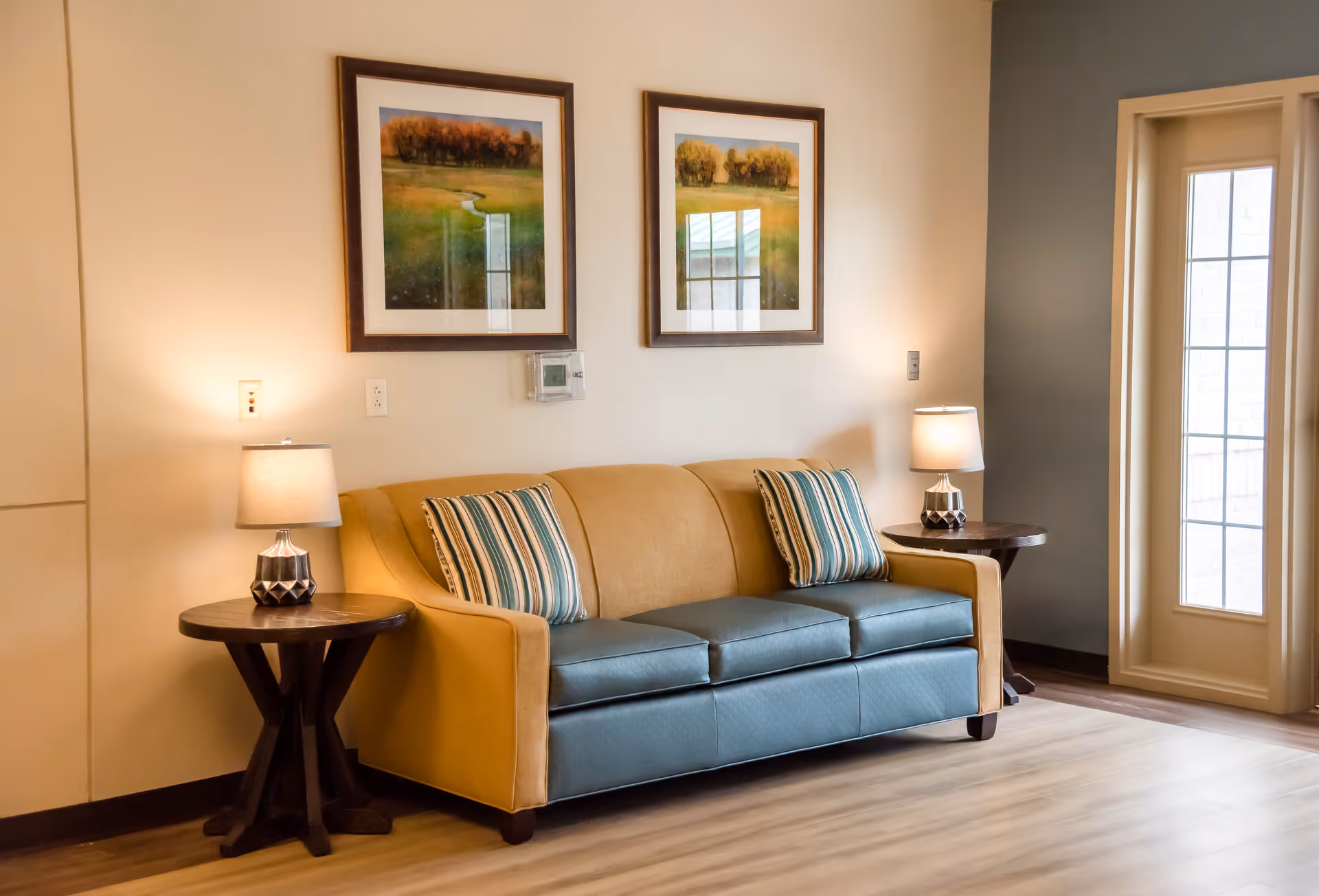 A cozy living room area with a two-tone sofa featuring mustard yellow sides and blue cushions, adorned with two striped pillows. On each side of the sofa, there are dark wooden side tables with table lamps. Above the sofa, two framed landscape paintings hang on the wall. To the right, there is a tall window or door with multiple glass panes allowing natural light to enter the room. The floor is light wood, and the walls are painted in neutral tones.