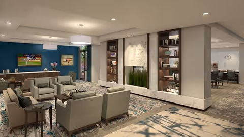 A spacious and modern senior living common area with comfortable armchairs arranged around a coffee table with a chessboard. The room features built-in shelves with decorative items, a large flat-screen TV mounted on a blue accent wall, and a dining area visible in the background. The space is well-lit with ceiling lights and pendant lamps.