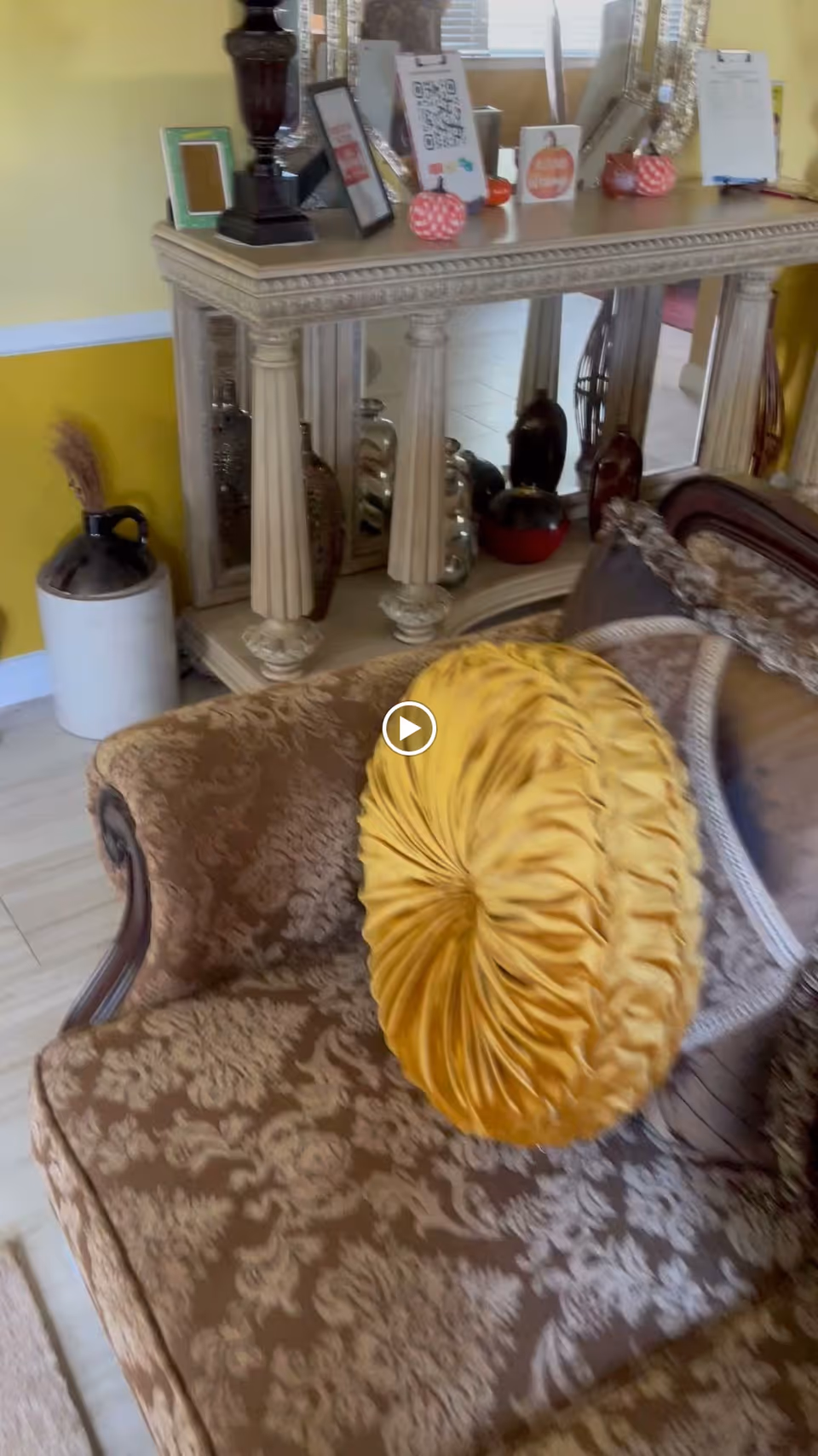 A cozy living room area with a patterned brown sofa adorned with a round yellow cushion. Behind the sofa is an ornate console table with decorative columns and a large mirror. The table holds various small decorative items, picture frames, and a lamp. The walls are painted yellow with white trim, and there is a large ceramic jug with dried plants on the floor next to the console table.