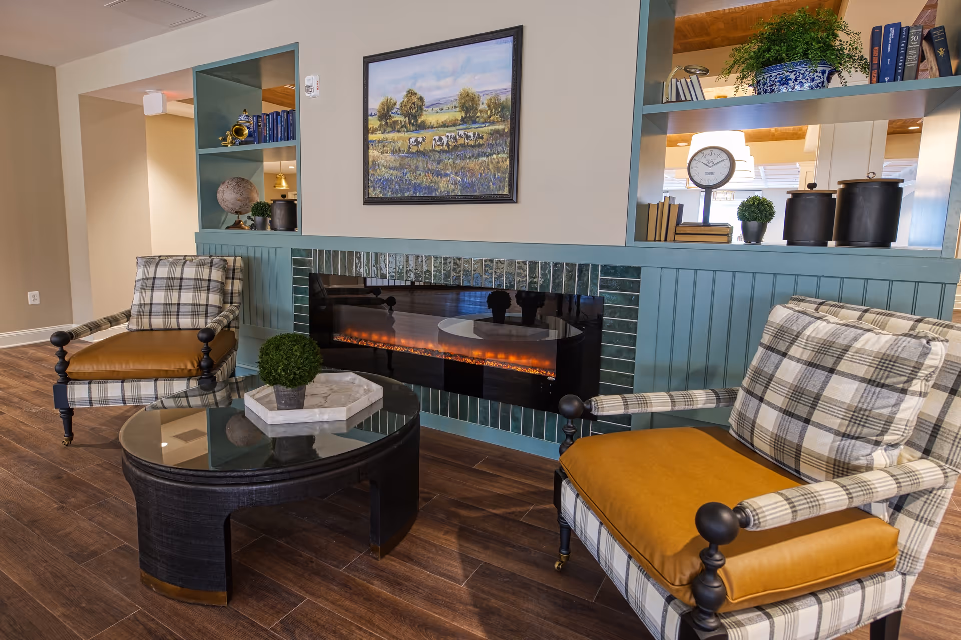 A cozy sitting area with two plaid armchairs featuring mustard-colored cushions, a black oval glass coffee table with a small potted plant, and a modern electric fireplace set into a teal paneled wall. Above the fireplace is a framed painting of a pastoral scene with cows in a field. Built-in shelves on either side of the fireplace hold books, decorative items, and plants. The floor is wood with a warm finish.