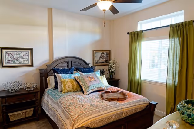 A cozy bedroom with a wooden bed frame and a colorful patterned bedspread. The bed is adorned with multiple decorative pillows. There are two framed pictures on the walls, a nightstand with decorative items, a window with green curtains, and a ceiling fan with a light fixture.