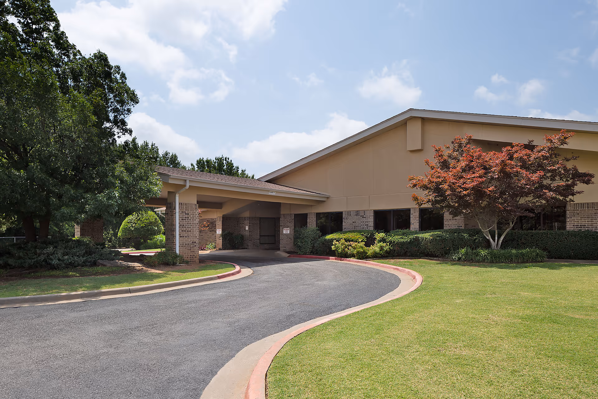 Exterior view of Brookwood Skilled Nursing & Therapy building entrance with a curved driveway, green lawn, trees, and shrubs under a partly cloudy sky.
