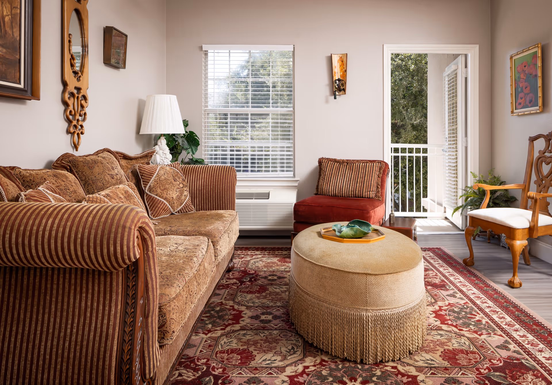 Cozy living room with a patterned sofa, ottoman coffee table, accent chairs, area rug, and an open door leading to a small balcony.