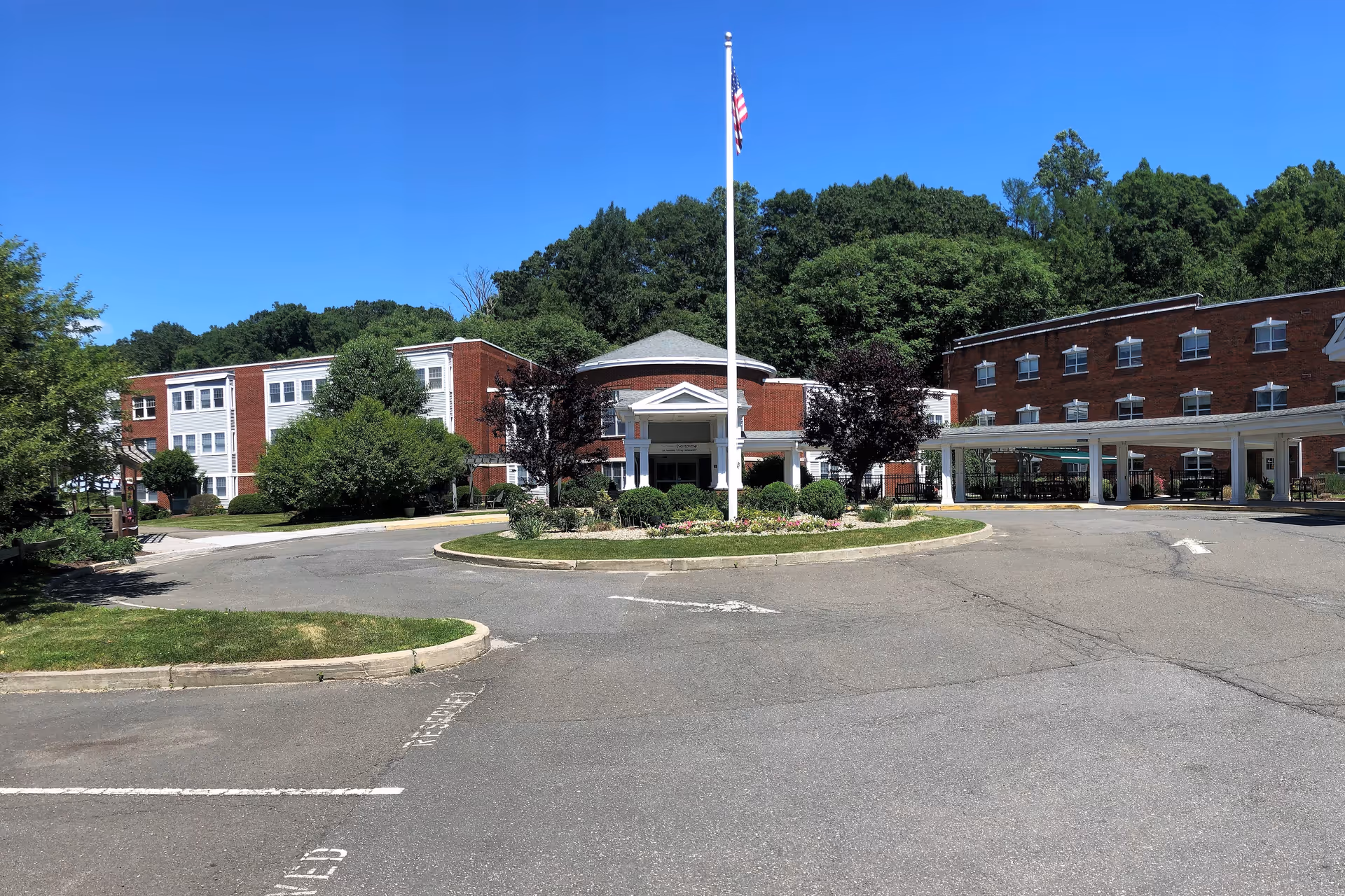 Front exterior view of The Cascades at Stone Bridge facility with a circular driveway, landscaped greenery, an American flag on a flagpole, and a clear blue sky.