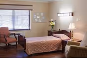 A cozy bedroom in a senior living facility with a single bed covered in a pink quilt, a wooden nightstand with a lamp and a vase of flowers, two chairs near a window with blinds, and framed pictures on the beige walls.