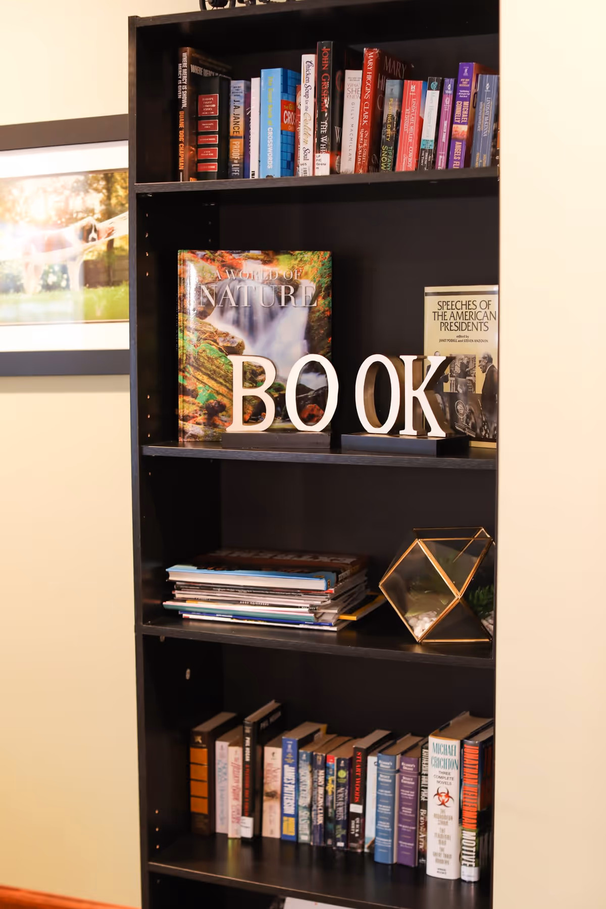 A black bookshelf filled with various books and magazines. On the middle shelf, there are decorative letters spelling out the word 'BOOK'. A framed picture is partially visible on the wall to the left of the bookshelf.