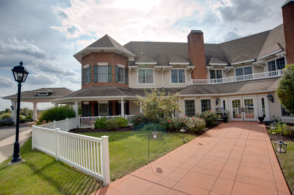 Front view of a two-story brick-and-siding senior living building with a red paved walkway, white picket fence, landscaped shrubs, and a lamppost.