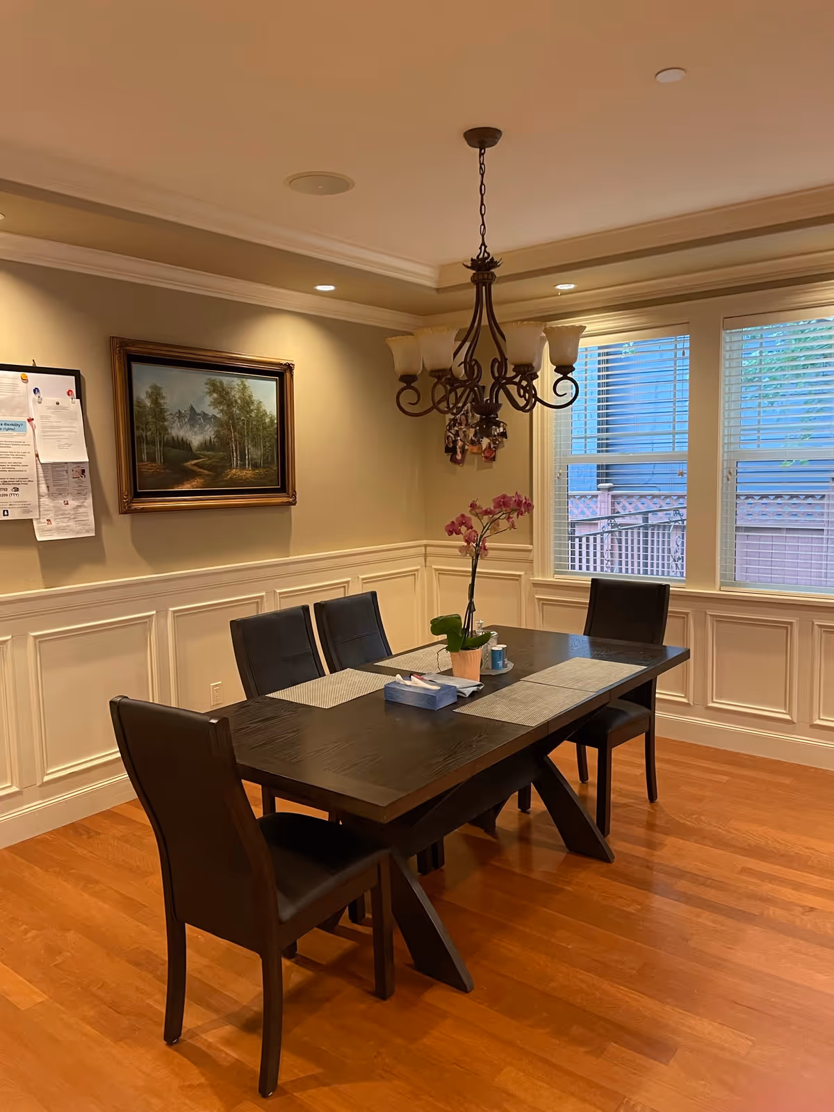 Dining room with a dark wood table and chairs, a hanging chandelier, a framed painting, and windows with blinds.