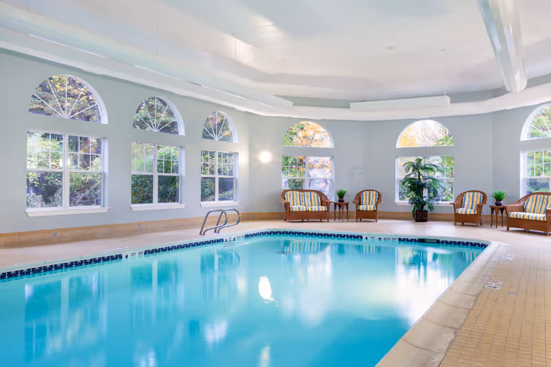 Bright indoor swimming pool with arched windows, lounge chairs, and potted plants.