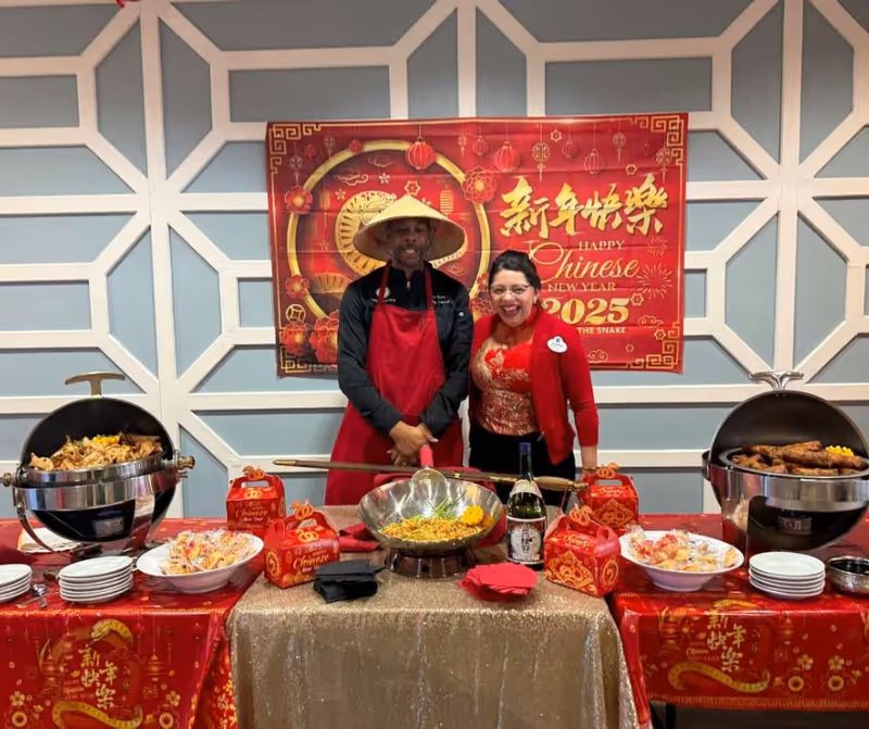 Two people standing behind a buffet table decorated for Chinese New Year 2025. The man on the left is wearing a traditional conical hat and a red apron, while the woman on the right is wearing a red and gold outfit. The table is covered with red and gold decorations, food trays with various dishes, and red gift boxes. A red banner with Chinese characters and 'Happy Chinese New Year 2025 Year of the Snake' is displayed on the wall behind them.