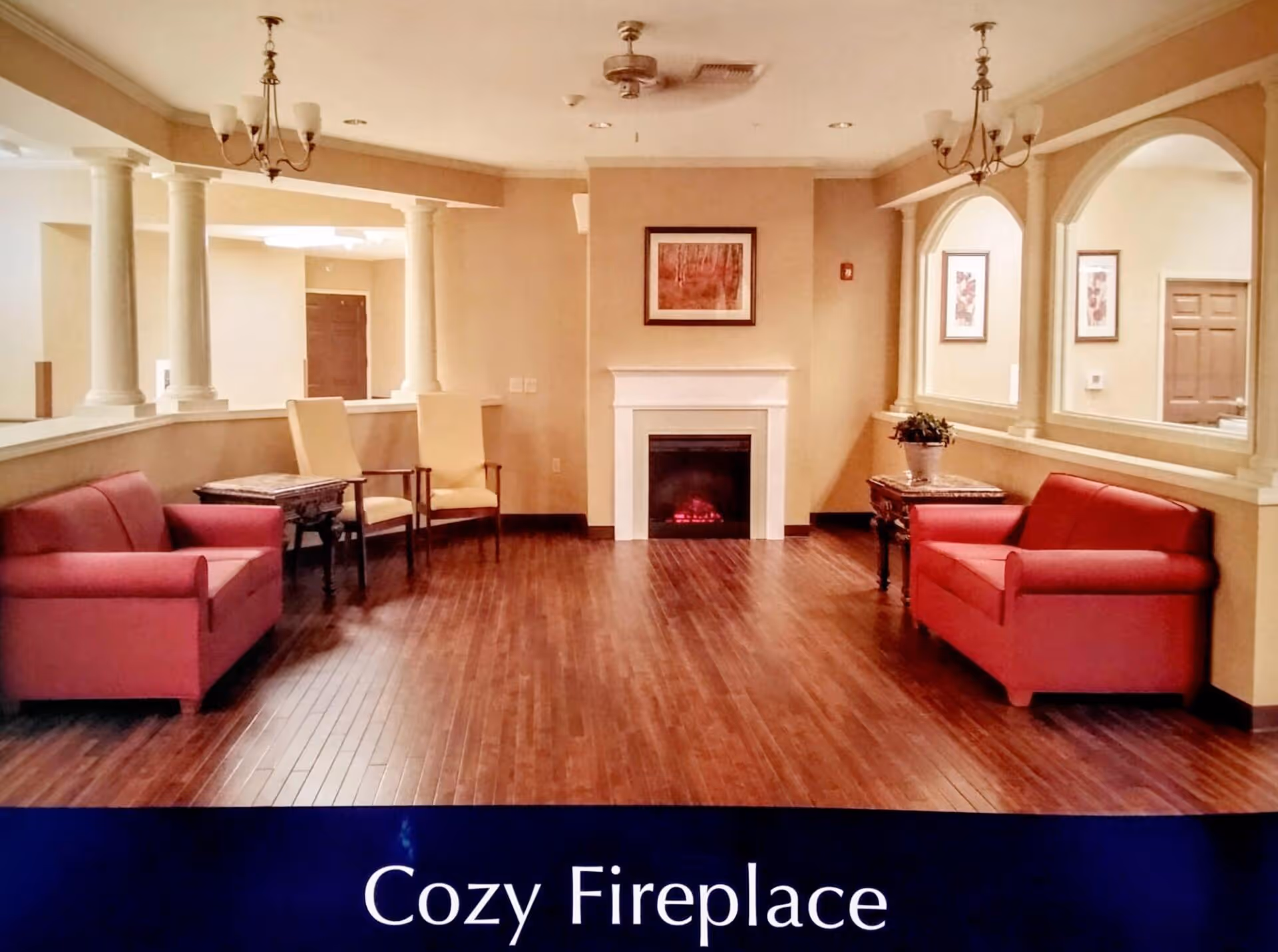 A cozy living room area with two red sofas facing each other, two beige chairs, a small table, a fireplace in the center with a framed picture above it, wooden flooring, and warm beige walls with decorative columns and arches.