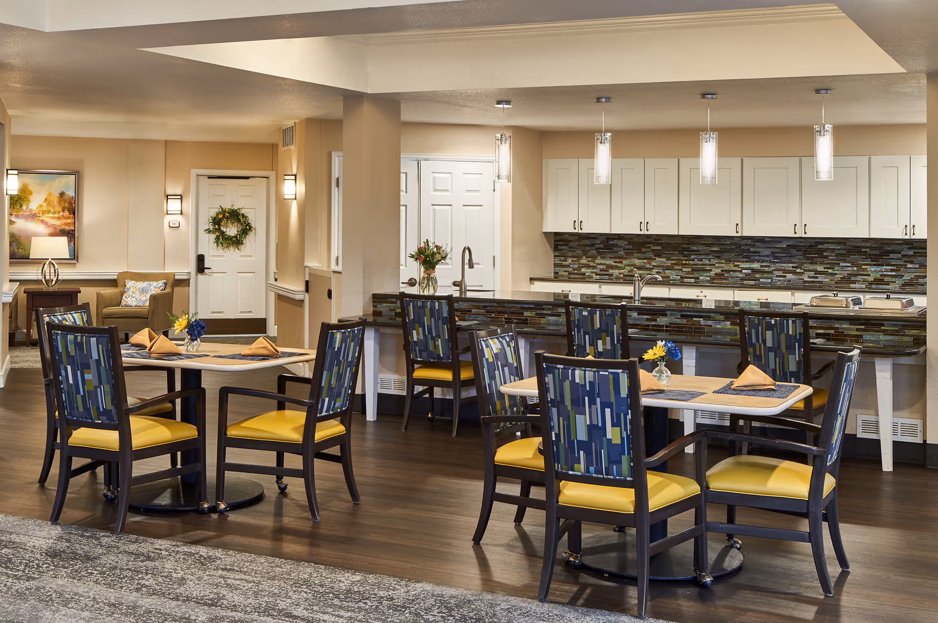 A bright and modern dining area in an assisted living facility featuring two square tables each surrounded by four chairs with yellow cushions and patterned backs. The tables are set with placemats, folded napkins, and small flower vases. In the background, there is a kitchen area with white cabinets, a tiled backsplash, and pendant lights hanging from the ceiling. A seating area with an armchair, side table, lamp, and framed artwork is visible near a white door decorated with a green wreath.
