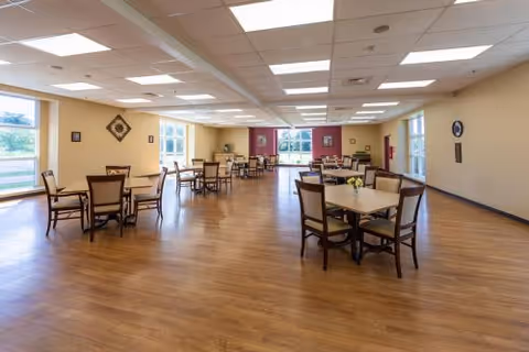 A spacious dining room with several square tables and chairs arranged neatly on a wooden floor. The room has large windows letting in natural light, light-colored walls, and a ceiling with recessed lighting panels. Some wall decorations are visible, and the room appears clean and well-maintained.