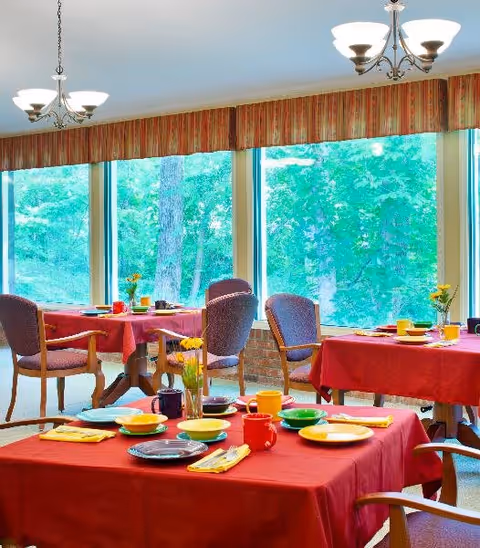 Dining room with several tables covered in red tablecloths, each set with colorful plates, cups, and yellow napkins. The room has large windows showing green trees outside and is lit by ceiling light fixtures.