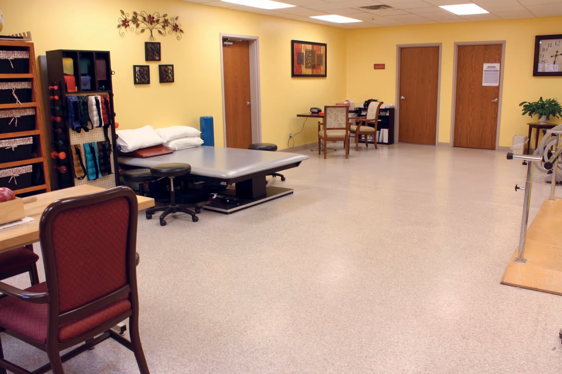 A spacious therapy or activity room with yellow walls and a light-colored floor. The room contains a large padded table with stools around it, a rack with various exercise bands and small weights, a couple of chairs and a desk in the background, and a clock on the wall. There are three wooden doors along the back wall and some decorative wall art.
